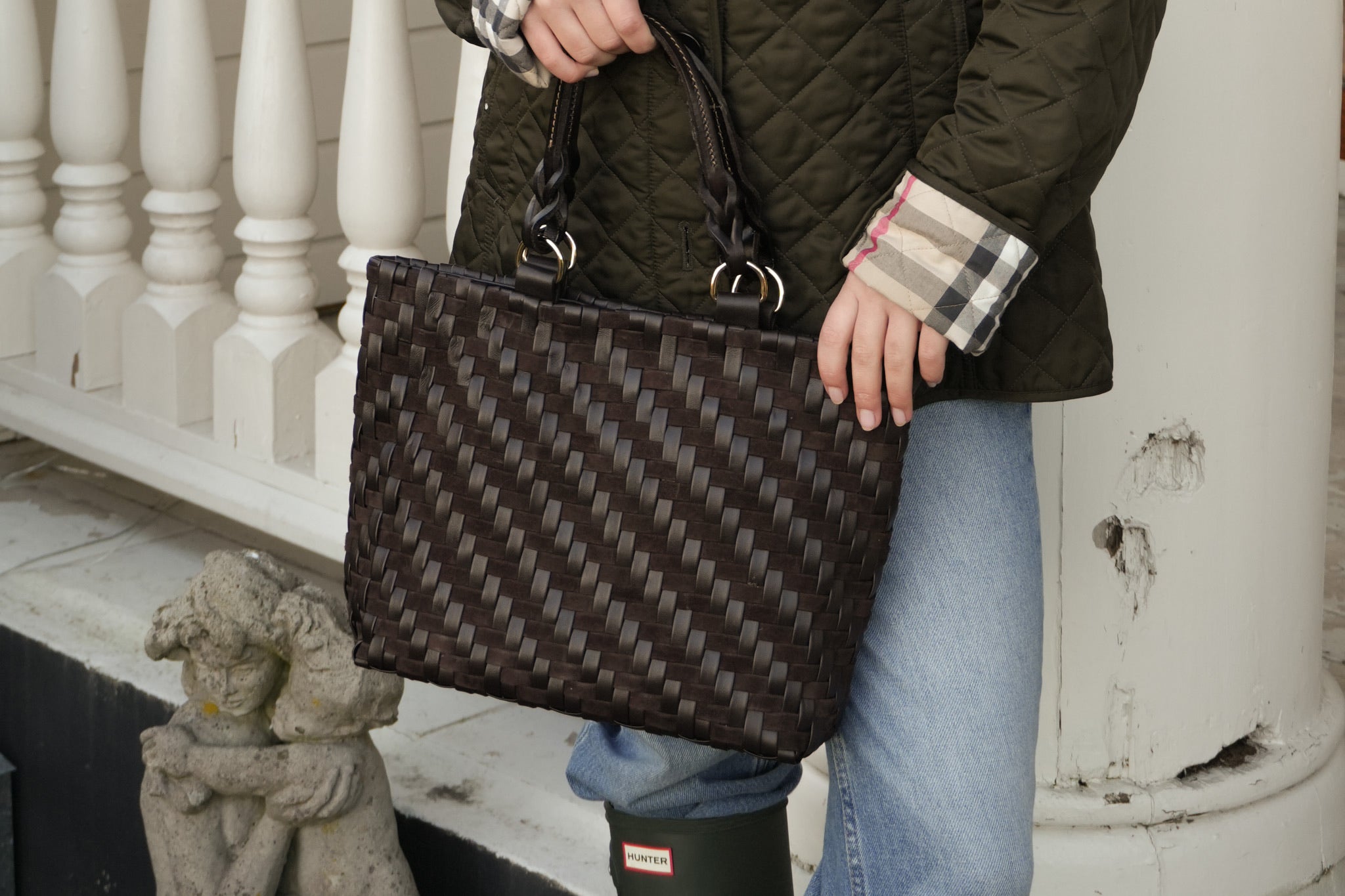 Person i grønn vattert jakke og jeans med Safari Lady Royal Chocolate, en håndflettet mørk brun skinnveske, på veranda.
