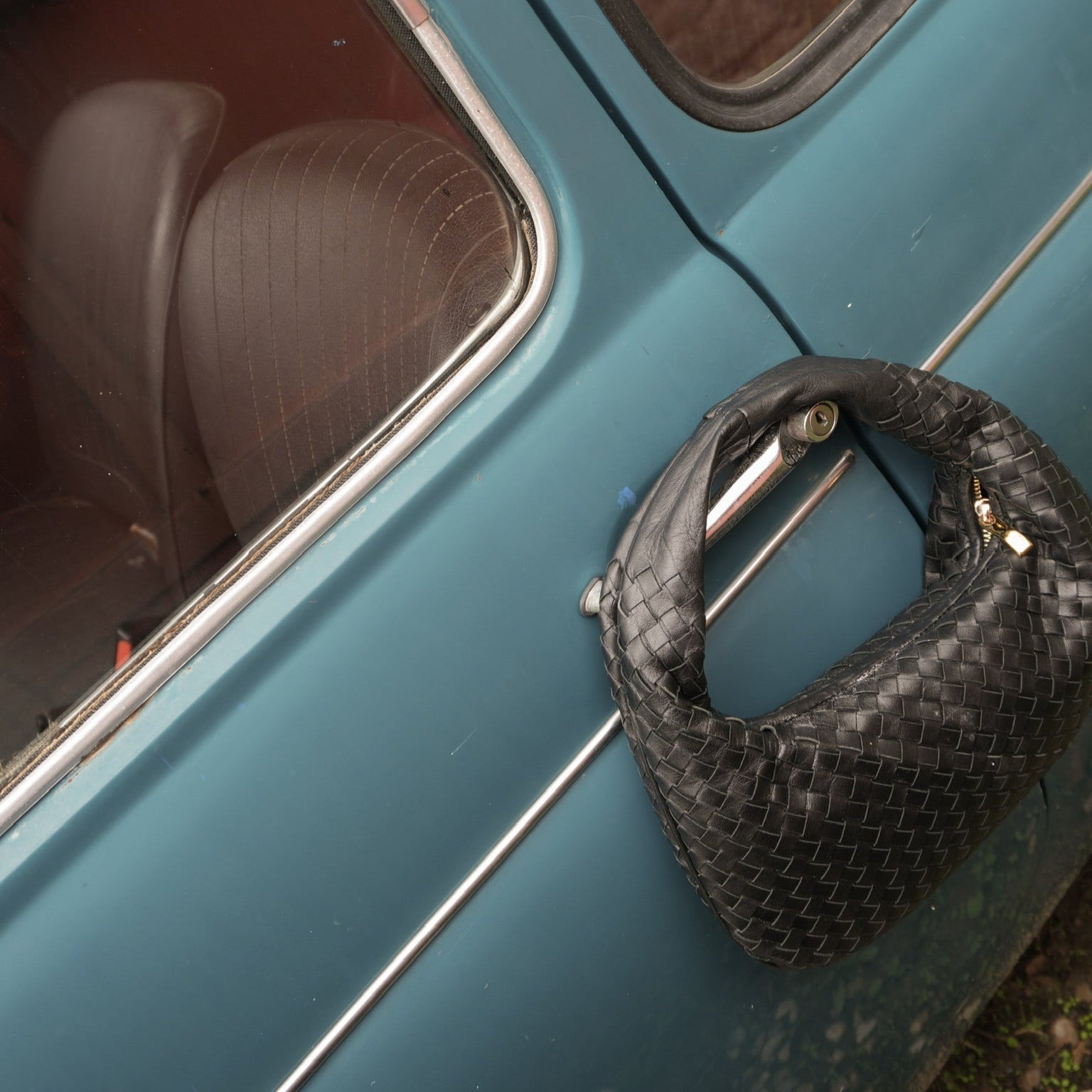 Sort flettet Isola Bella-veske henger elegant på døren til en vintage blå bil.