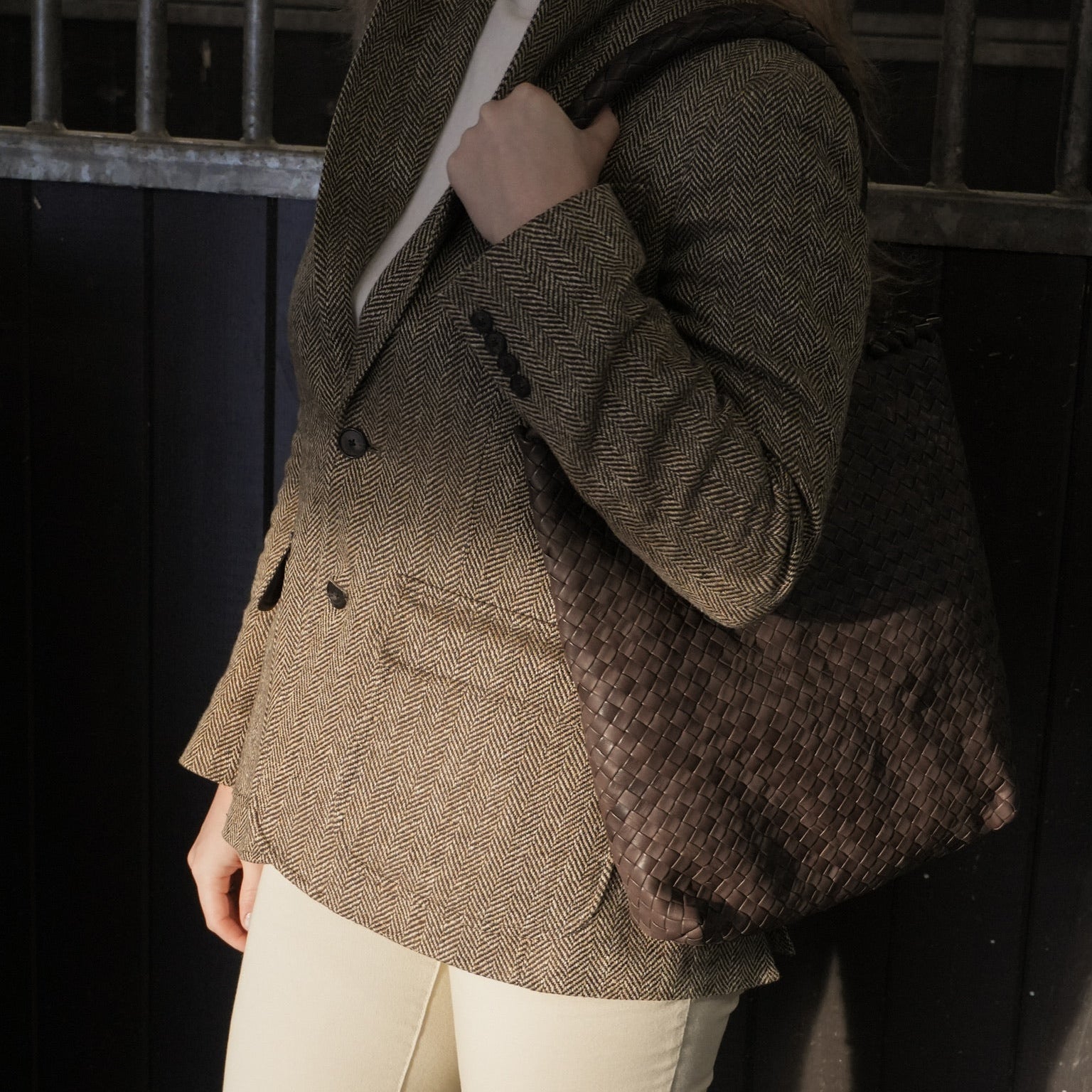 Kvinne i tweedjakke bærer Ribichini Woodstock Babe Brown, en brun håndflettet toteveske, i et stallmiljø med mørke vegger.