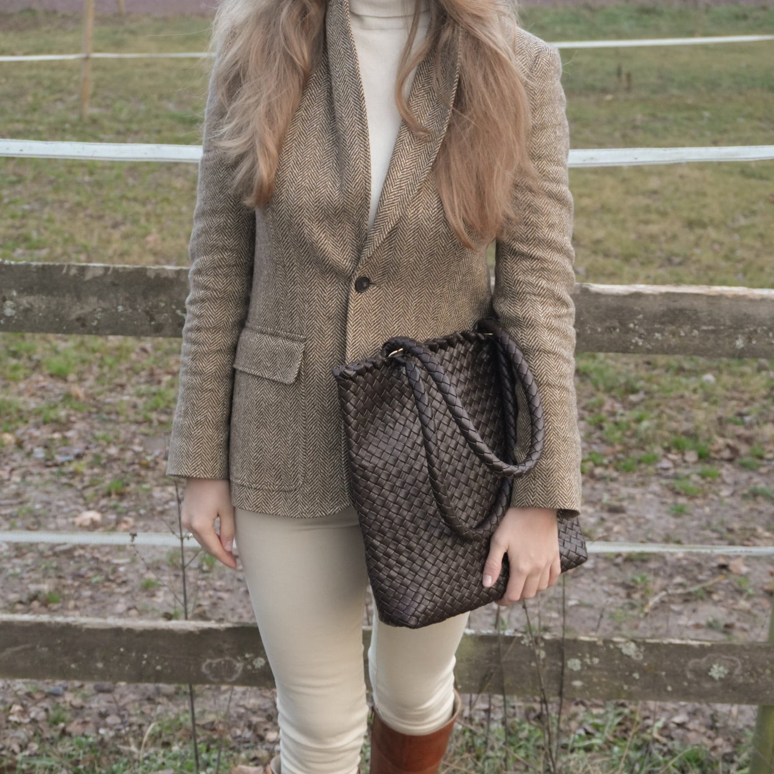Kvinne i tweedjakke holder Ribichini Woodstock Babe Chocolate, en brun håndflettet toteveske, foran et gjerde ute i naturen.