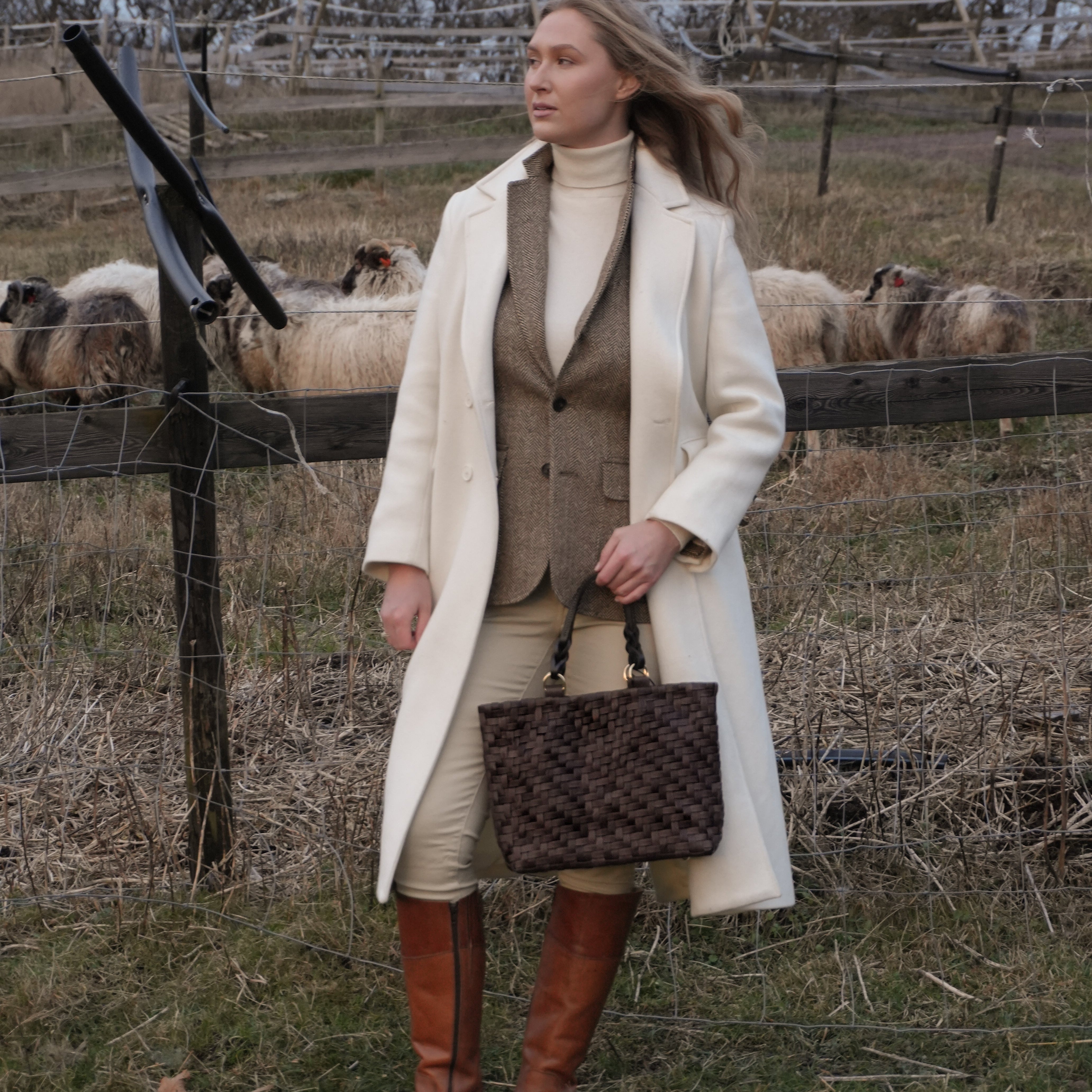 Kvinne i lys kåpe og brune støvler holder Ribichini Safari Lady i brun flettet skinn ved en saueinnhegning i naturen.