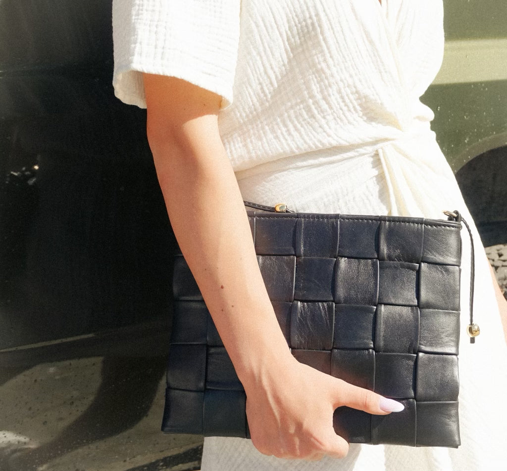 Square Clutch i flettet svart skinn, båret elegant mot en hvit kjole i sollys for et tidløst og moderne uttrykk.