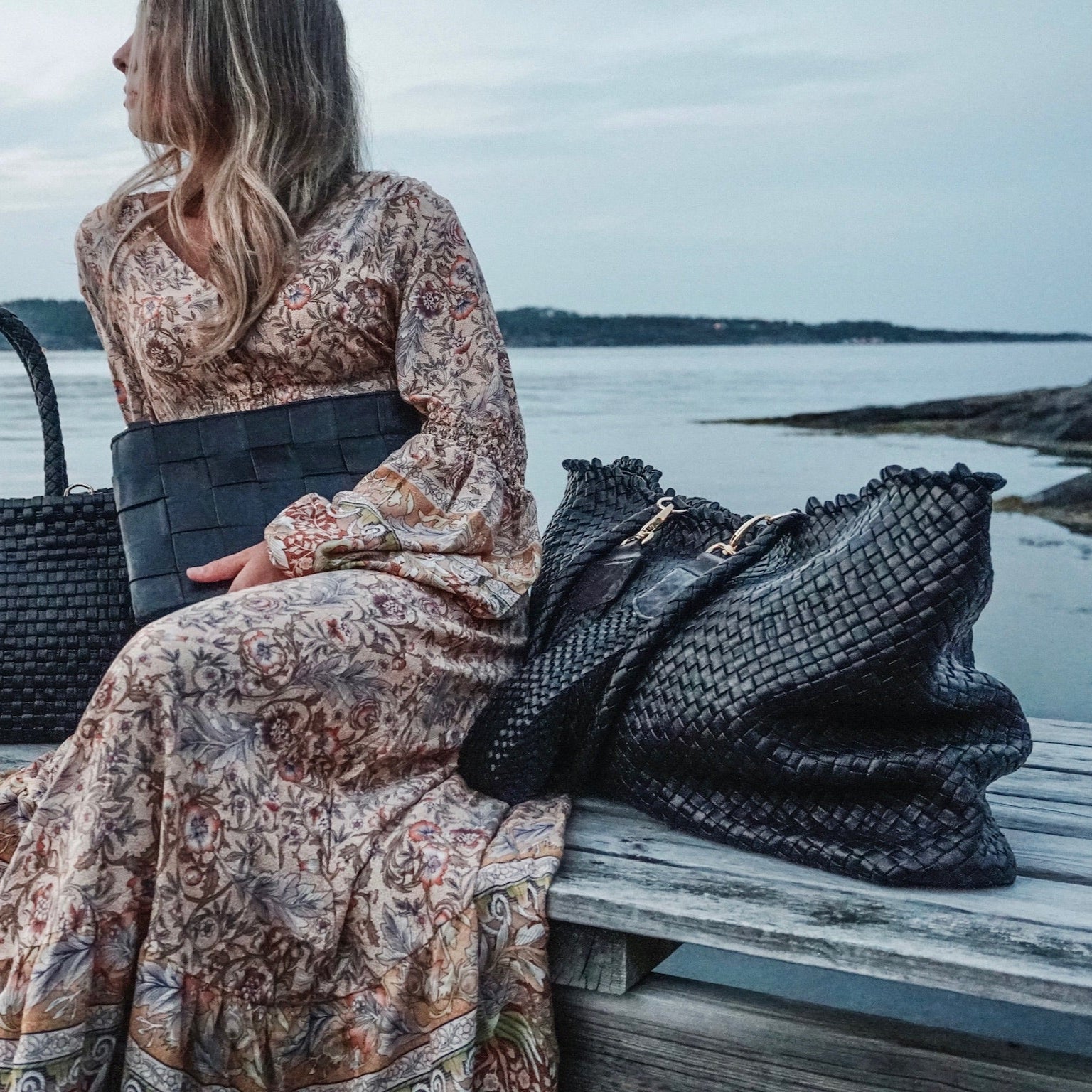 Kvinne ved sjøen med Ribichini Woodstock Weekend i sort håndflettet skinn – eksklusiv veske med tidløs eleganse.