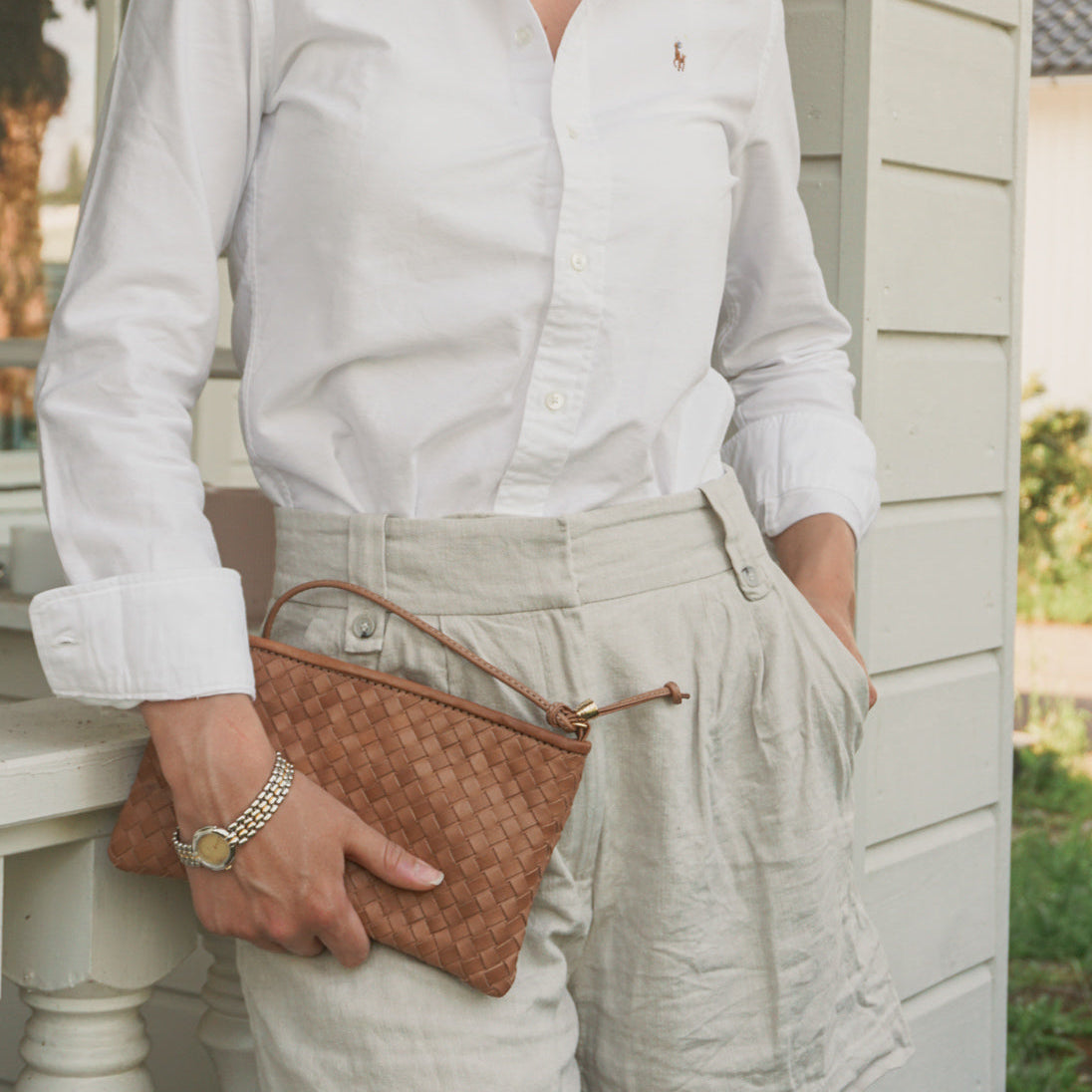 Kvinne i hvit skjorte og linshorts holder Ribichini Camper Clutch i cognac flettet skinn – elegant og tidløs.