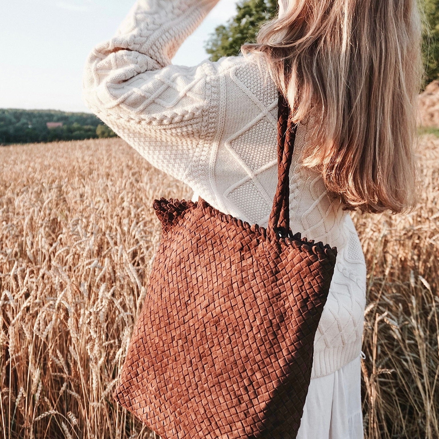 Kvinne i åker med Ribichini Woodstock veske i brunt semsket skinn, håndlaget i flettet design.