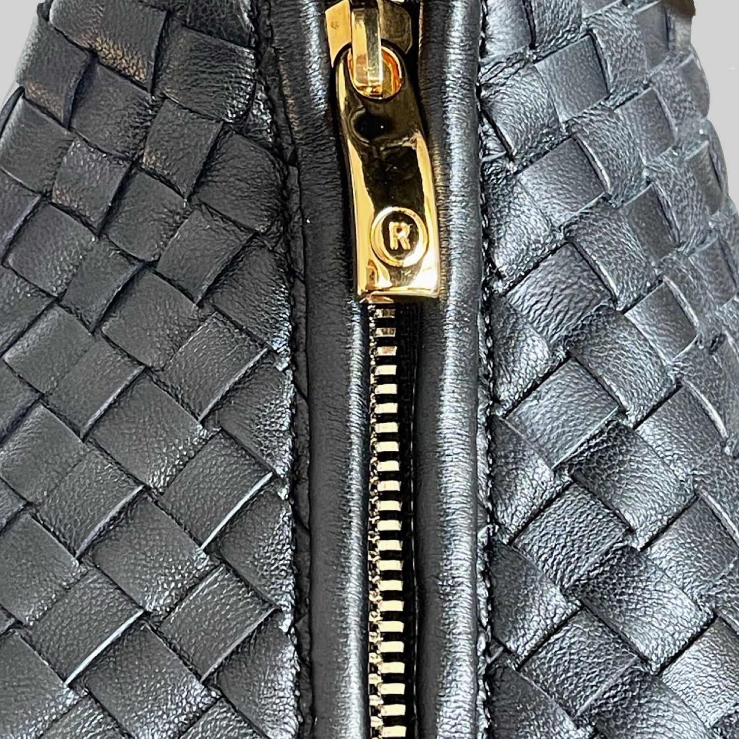 Detaljert bilde av Ribichini-veskens glidelås i gull med merkevarelogoen R, mot flettet svart lammeskinn.