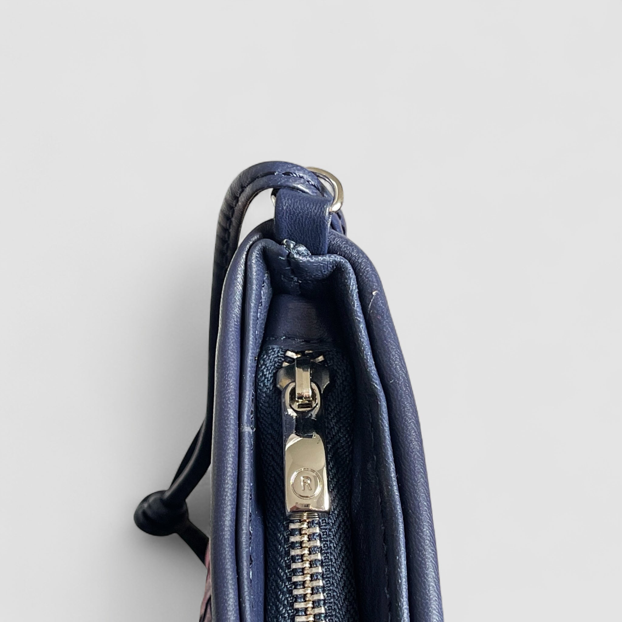 Detaljbilde av glidelås på navyblå Ribichini clutch – sølvfarget med preget R-logo og flettet skinnkant.