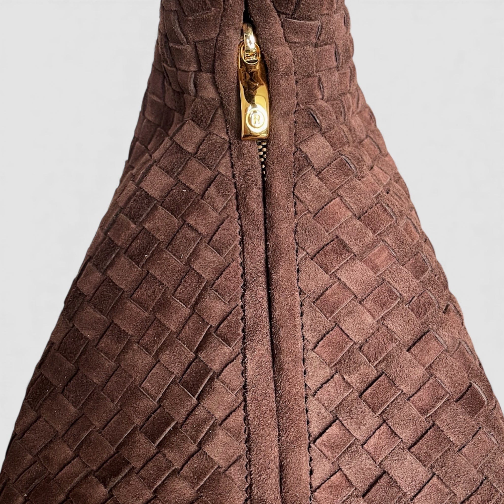 Detaljert bilde av Ribichini-veskens glidelås i gull med merkevarelogoen R, mot flettet semsket lammeskinn.