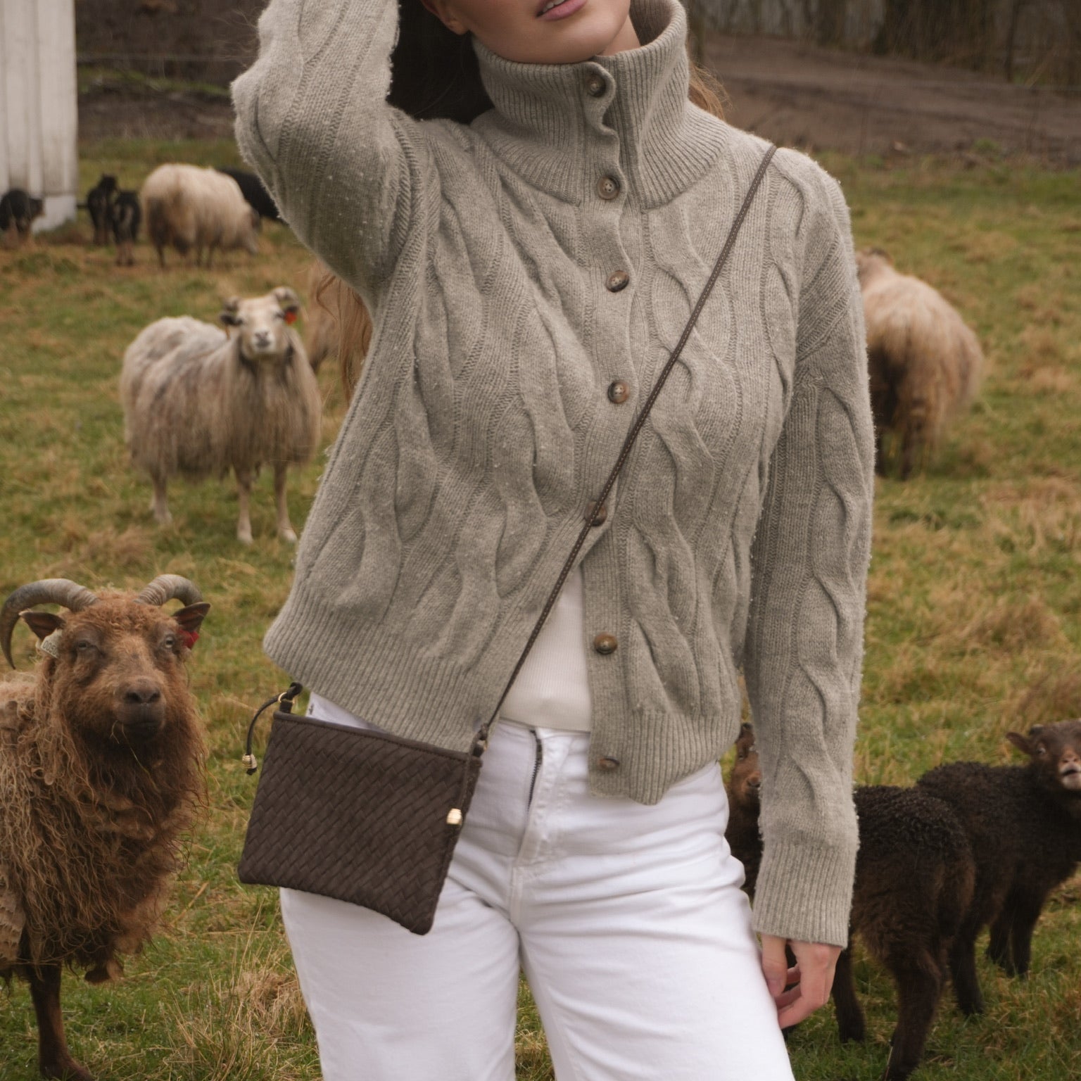 Kvinne i lys grå kabelstrikk bærer Ribichini Camper Clean Clutch Brown, en håndflettet clutch i mørkebrunt lammeskinn med lang skulderrem, ute på gresset blant sauer.