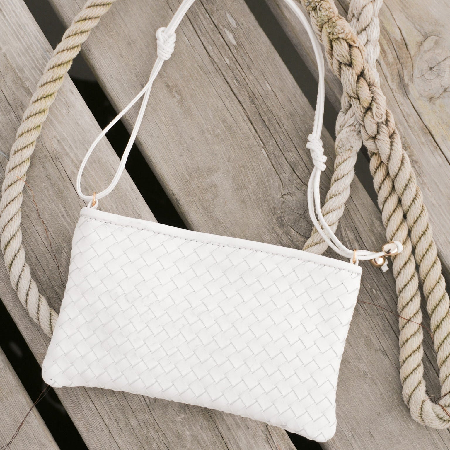 Ribichini Camper Clean Clutch White håndflettet skinnveske med justerbar hvit skulderrem, fotografert på en trebrygge med tau i bakgrunnen.