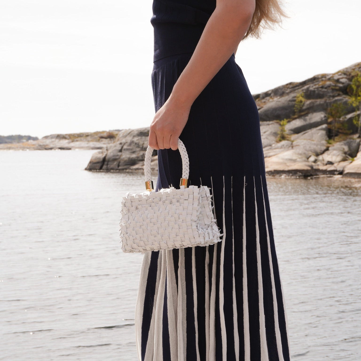 Ribichini Ribimini White håndflettet skinnveske med flettet håndtak, båret ved vannet og stylet med mørk blå og hvit sommerkjole.