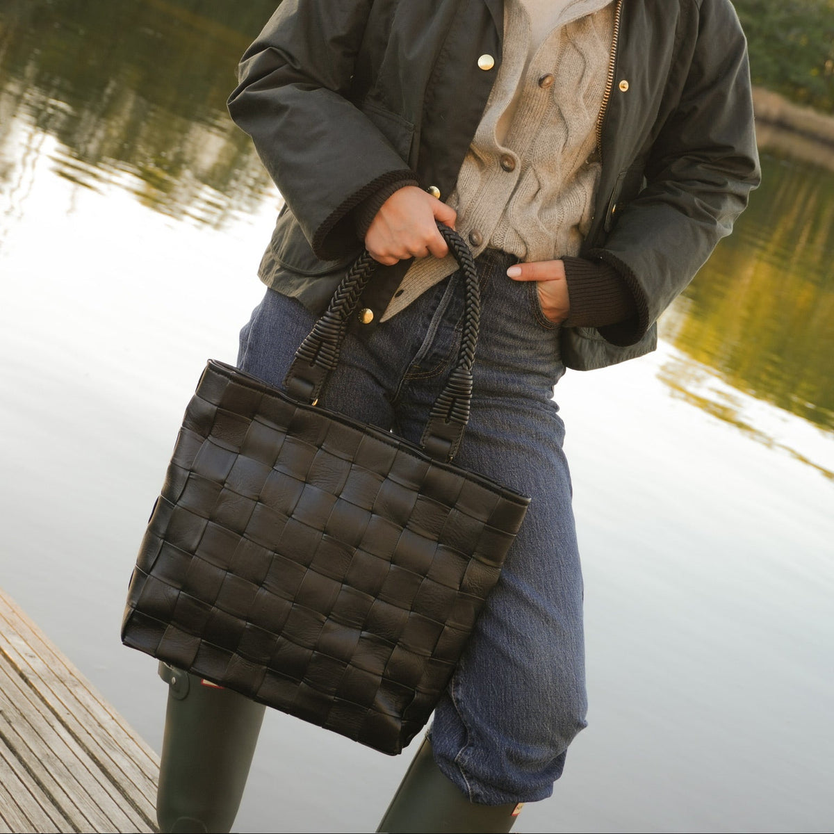 Modell med Ribichini Square Babe Black, en sort håndflettet veske i lammeskinn, stylet med høstklær ved vannet.