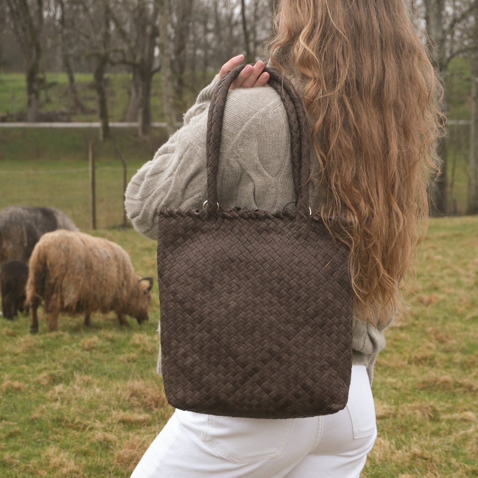 Kvinne med langt lyst hår bærer Ribichini Woodstock Babe Brown, en håndflettet skinnveske i mørkebrunt lammeskinn, over skulderen ute på gresset blant sauer.