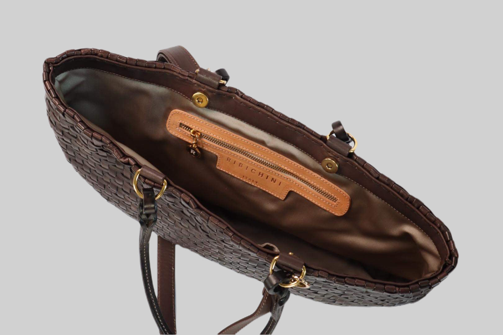 Bildet gir et innblikk i innsiden av en Ribichini Safari-veske i brunt. Den praktiske og organiserte vesken har en innvendig glidelåslomme i gullfarge og den ikoniske R-logoen til Ribichini.