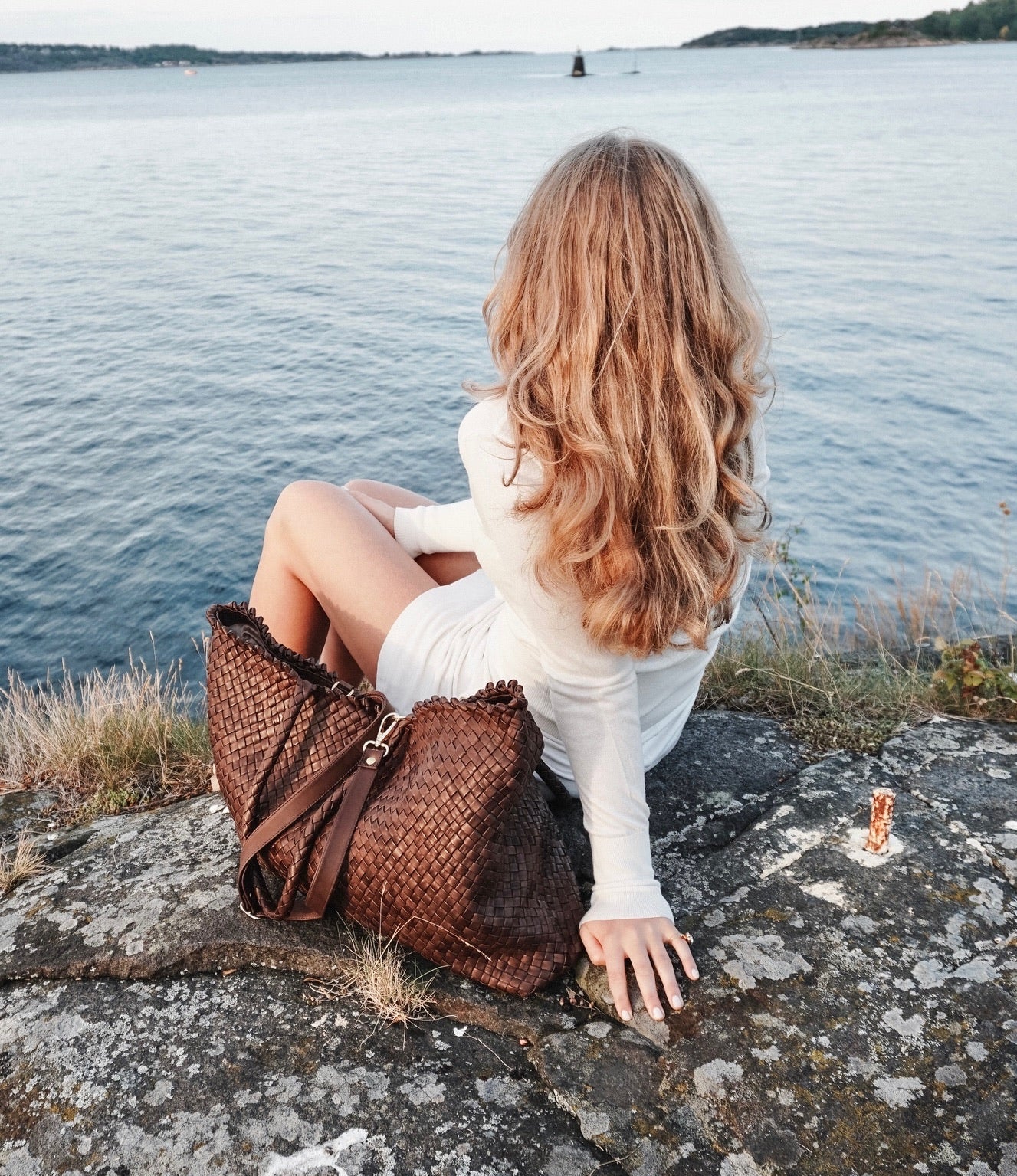 En ung kvinne ser utover havet ved siden av en Woodstock Weekend skinnveske i brun, preget av naturlig eleganse.