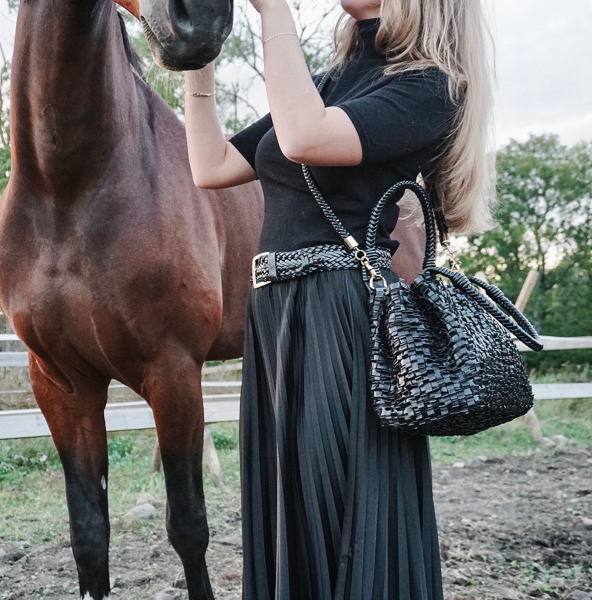 En ung kvinne står ved siden av en brun hest og klapper den. Med en Ribichini Loren skinnveske i sort på armen, utstråler hun tidløs eleganse.