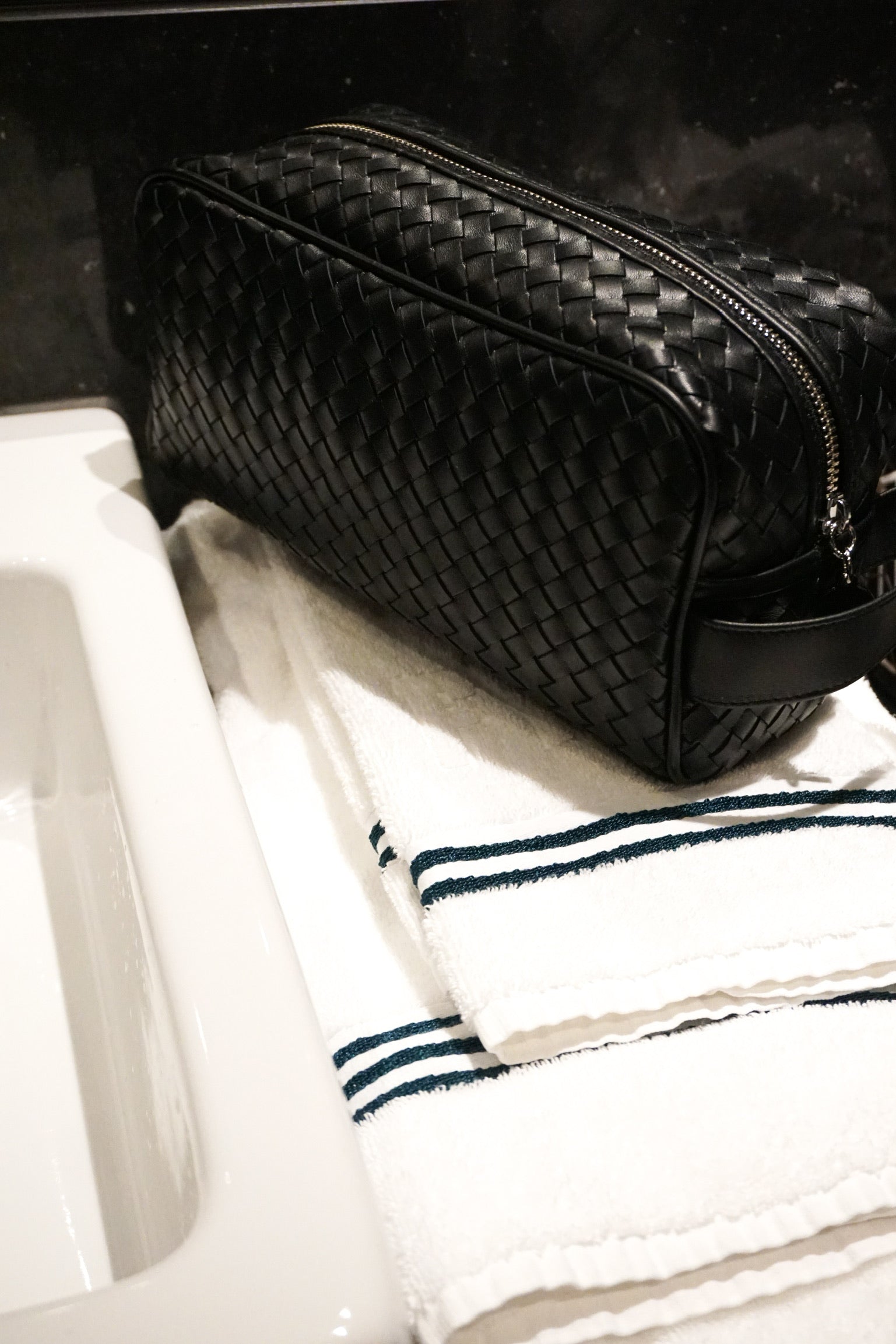 Ribichini Secrets toalettmappe i sort på hvit vaskeservant med blådetaljert håndkle. Elegant kontrast mellom teksturer og farger.