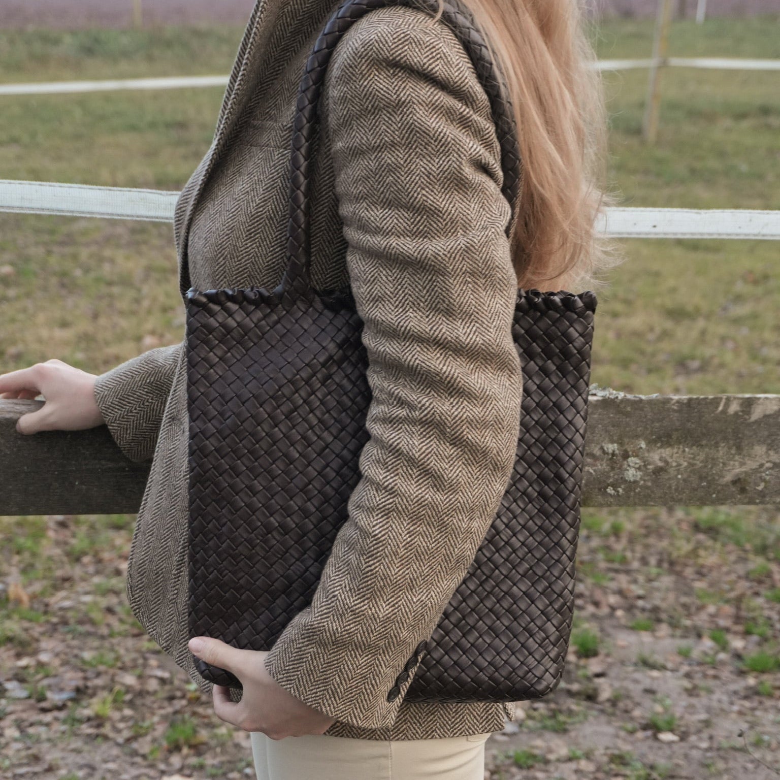 Kvinne i tweedjakke bærer Ribichini Woodstock Babe Chocolate, en brun håndflettet toteveske, ved et gjerde i naturen.