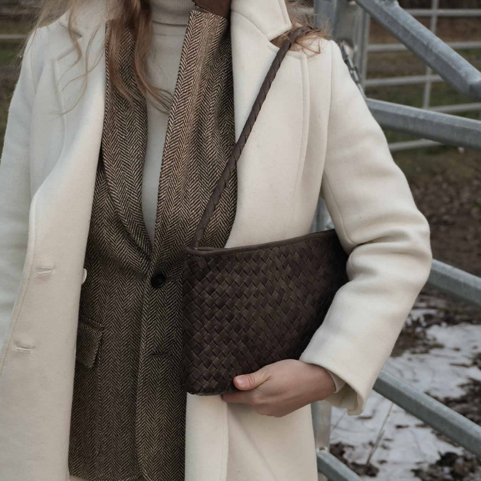 Kvinne i hvit kåpe og tweedjakke holder Ribichini Classica Brown, en brun håndflettet skulderveske, ved et gjerde ute i naturen.