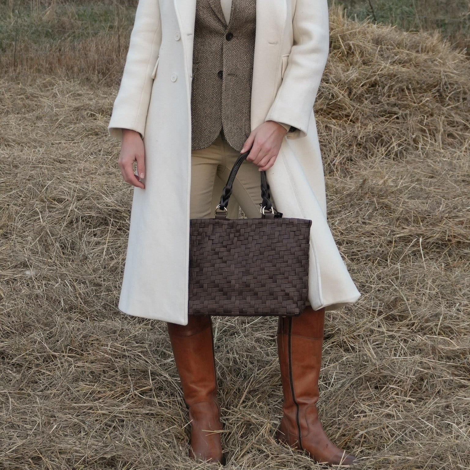 Kvinne i hvit kåpe holder Ribichini Safari Lady Brown, en brun håndflettet toteveske, mens hun står på et jorde foran en steinmur.