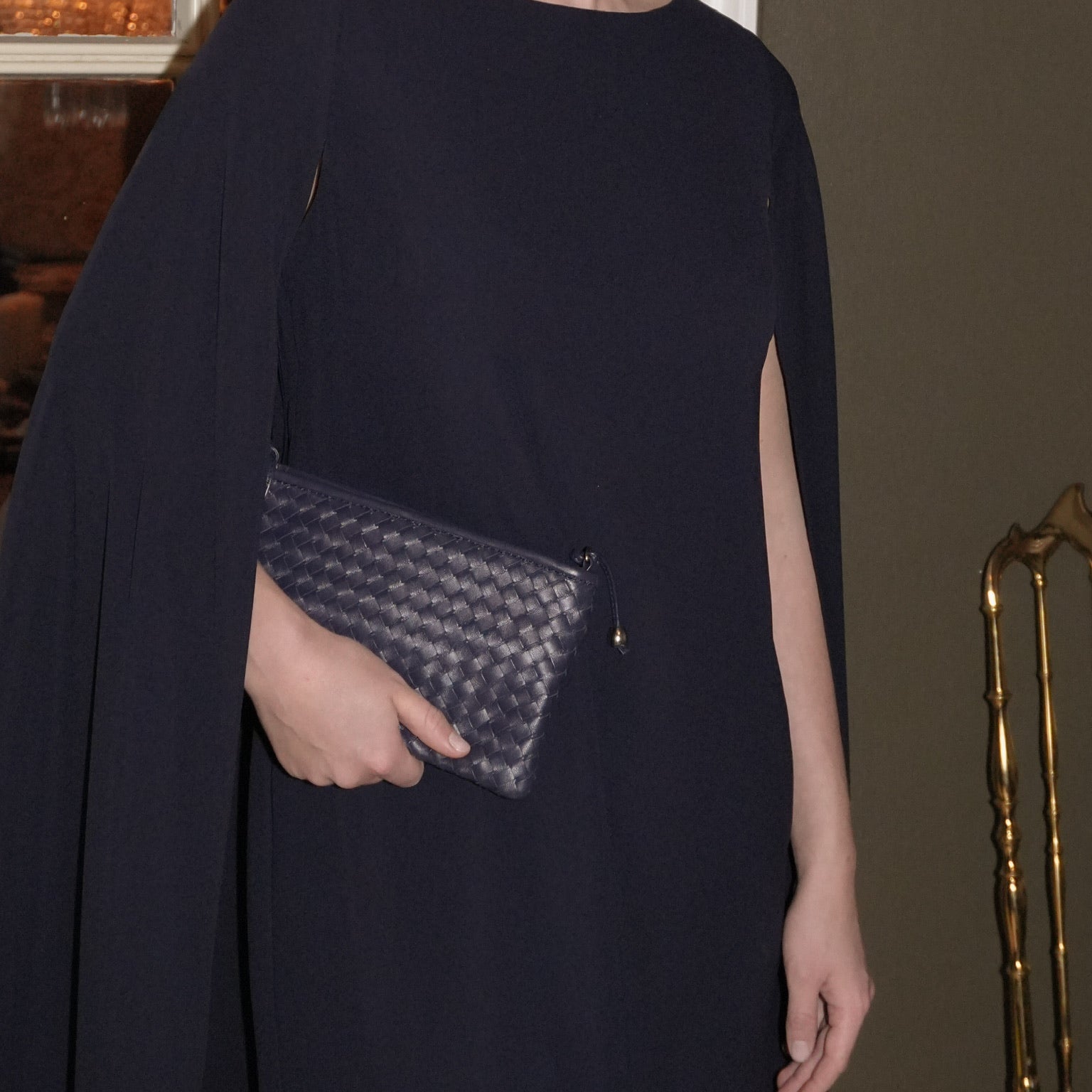 Kvinne i mørk kjole holder Ribichini Camper Clean Clutch Navy, en navy håndflettet clutch, i et elegant salongmiljø.