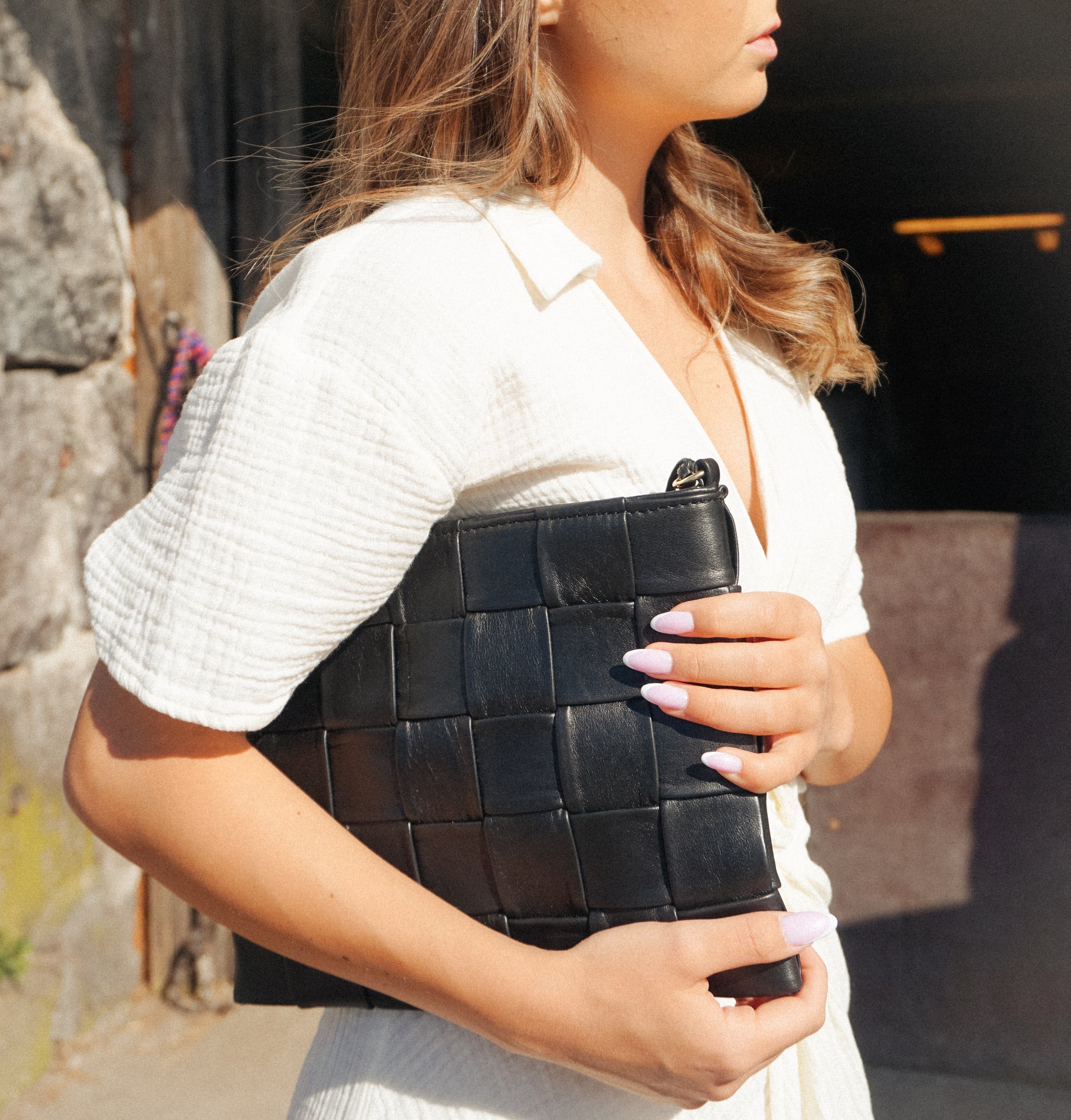 Square Clutch i flettet svart skinn, båret elegant mot en hvit kjole i sollys for et tidløst og moderne uttrykk.