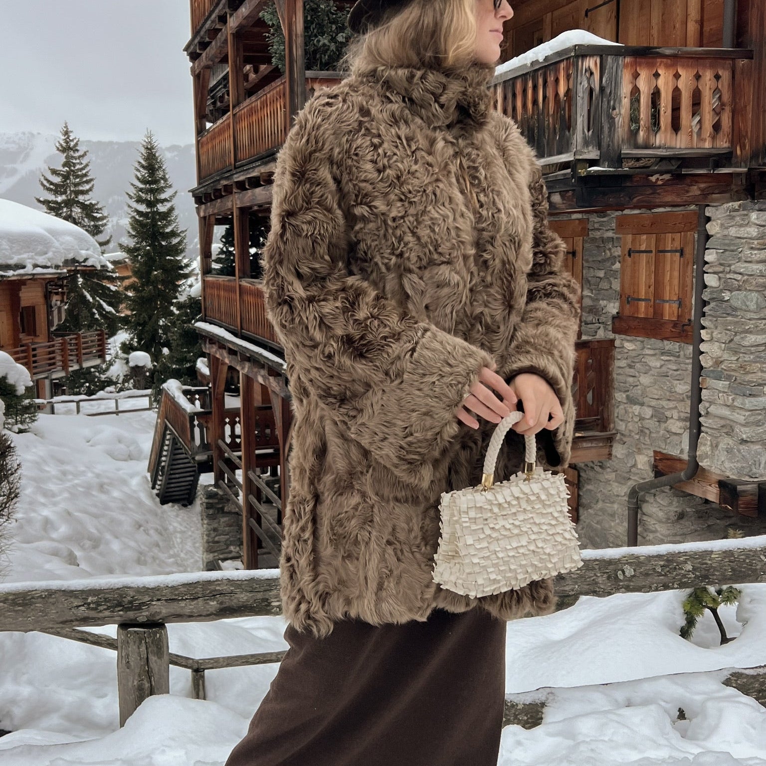 Ribimini Cream håndvevd kremfarget veske med strukturert overflate og korte håndtak, stylet med brun fuskepels og vinterantrekk foran alpehytte i snølandskap.