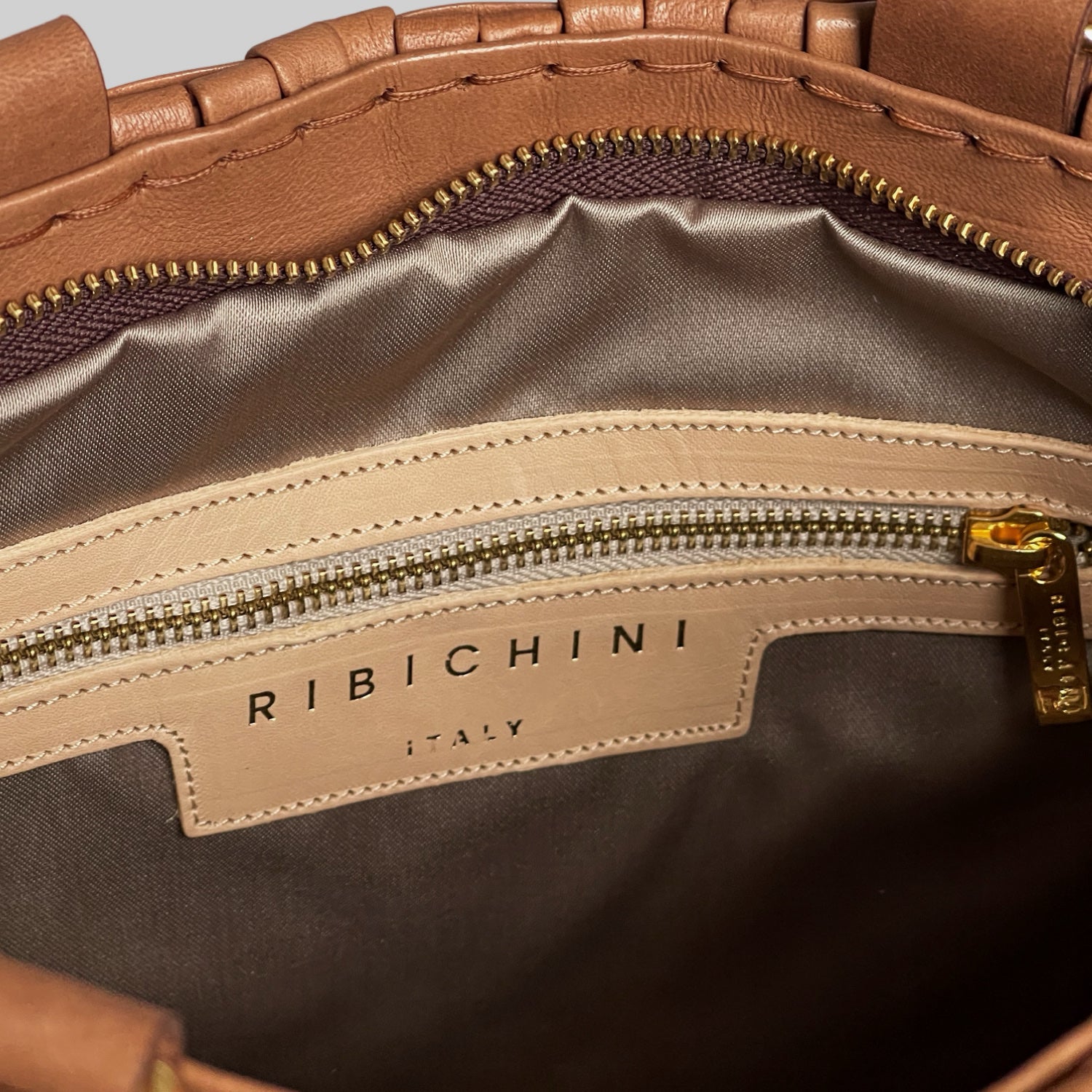 Bildet gir et glimt av innsiden av Ribichini Safari Lady-vesken i cognac, med en elegant glidelåslomme i gullfarge og den ikoniske R-logoen, som gir et preg av luksus og kvalitet.