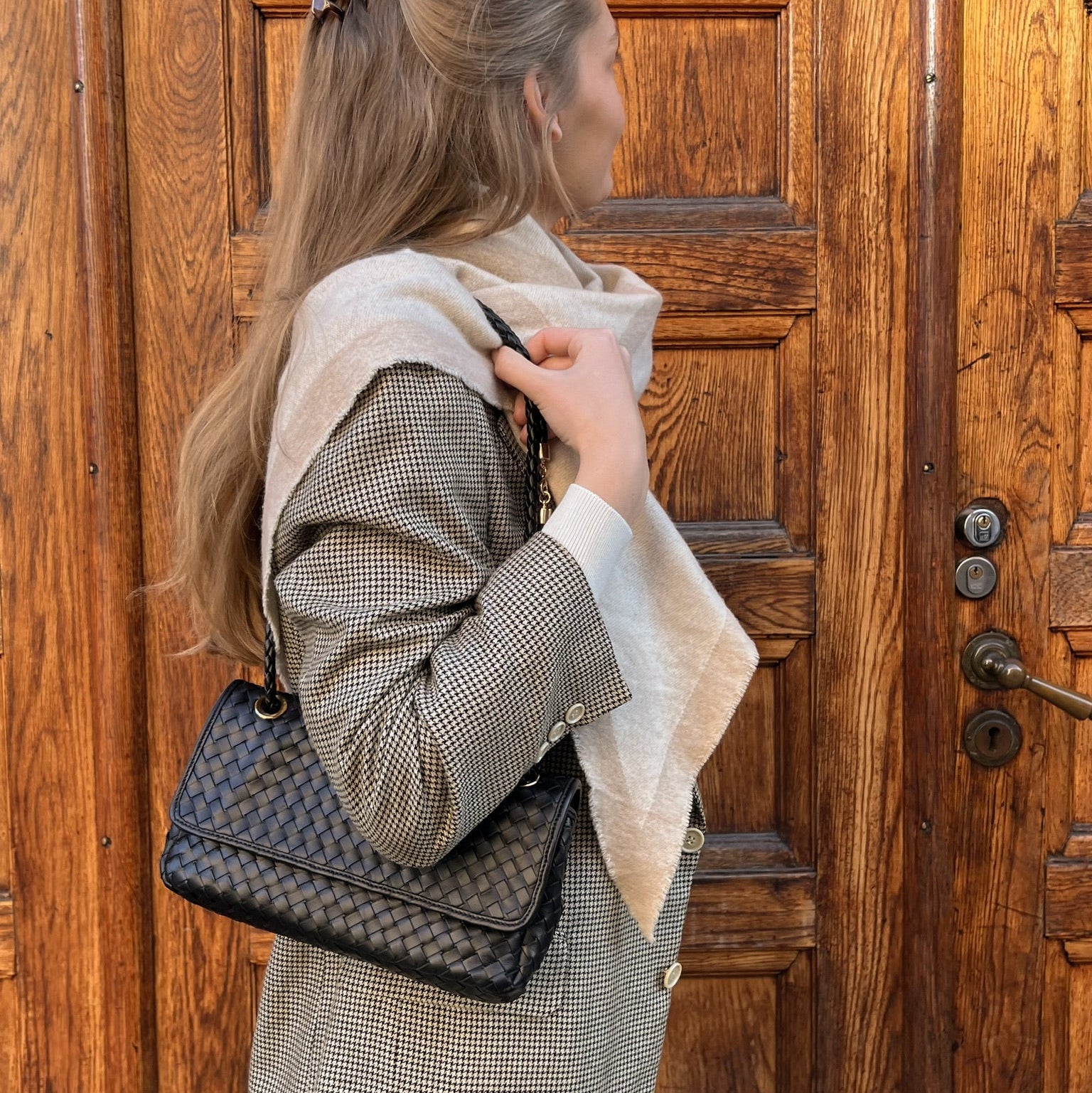 Klassisk Envelope veske i sort flettet skinn på skulderen, stylet med rutete blazer og beige ullskjerf foran tredør.