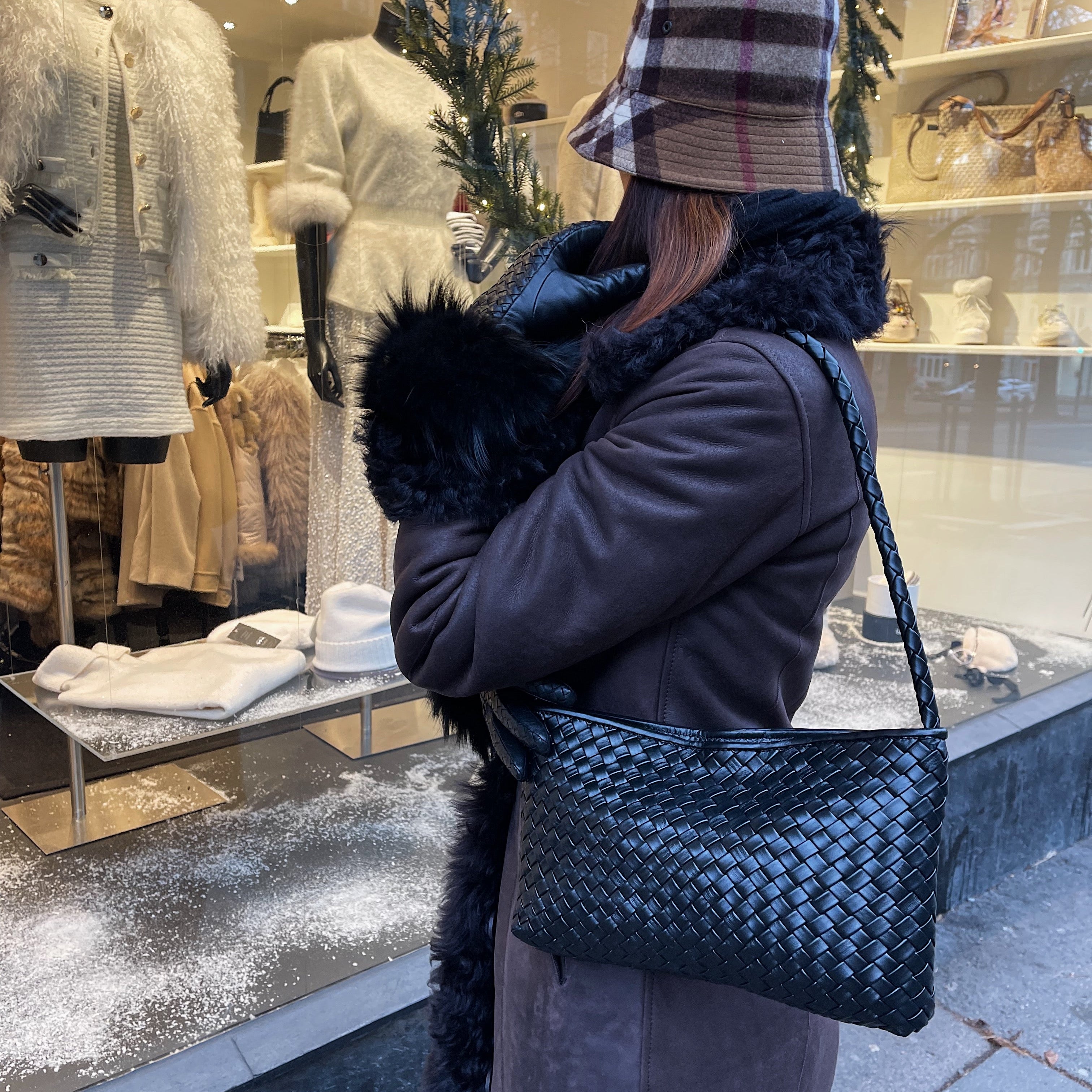 Person med brun kåpe og hatt bærer Ribichini Classica i sort håndflettet skinn foran et julepyntet butikksvindu.