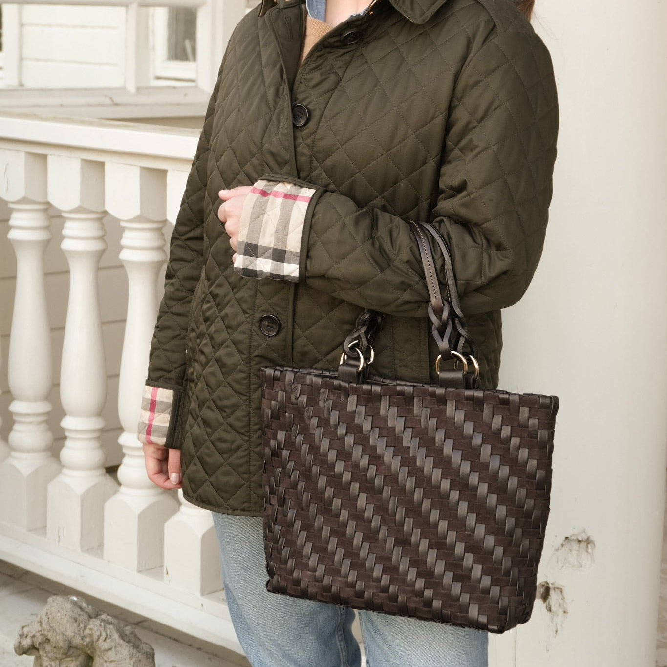 Person i grønn vattert jakke og jeans med Safari Lady Royal Chocolate, en håndflettet mørk brun skinnveske, på veranda.