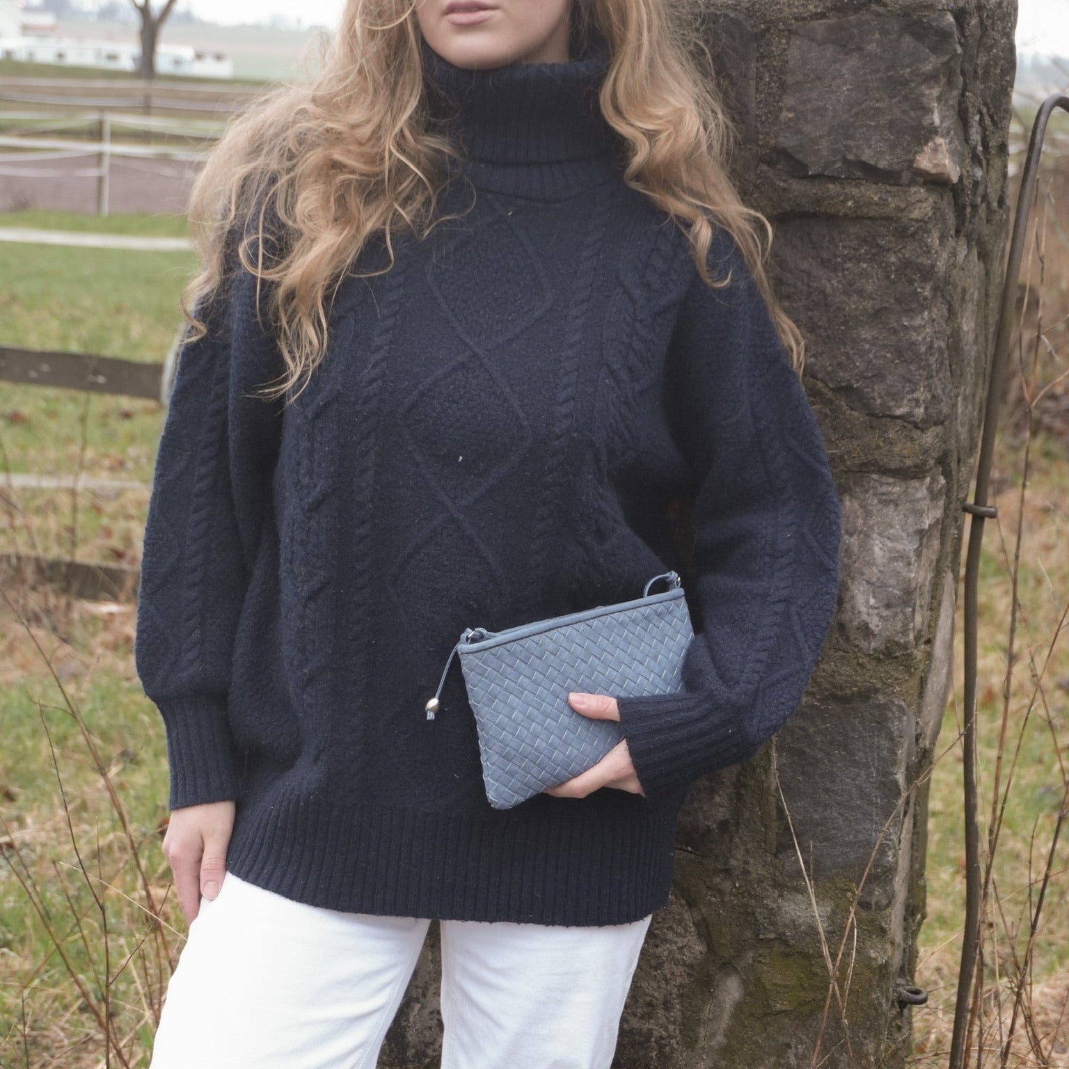 Kvinne i mørk marineblå strikkegenser holder Ribichini Camper Clean Clutch Jeans, en håndflettet clutch i blått lammeskinn.