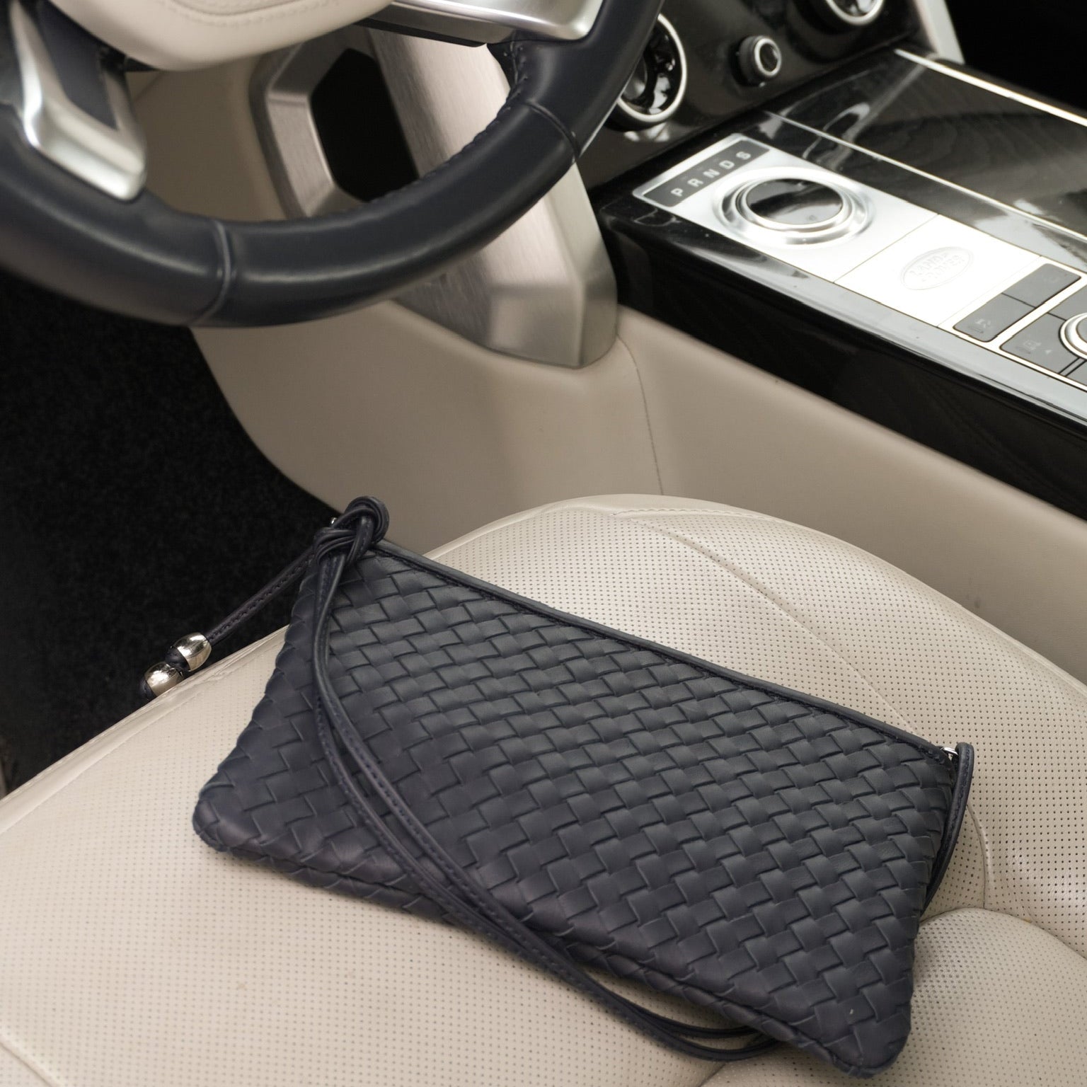 Ribichini Camper Clean Clutch Navy håndflettet skinnveske med skulderrem, plassert på et lyst bilsete i et eksklusivt bilinteriør.