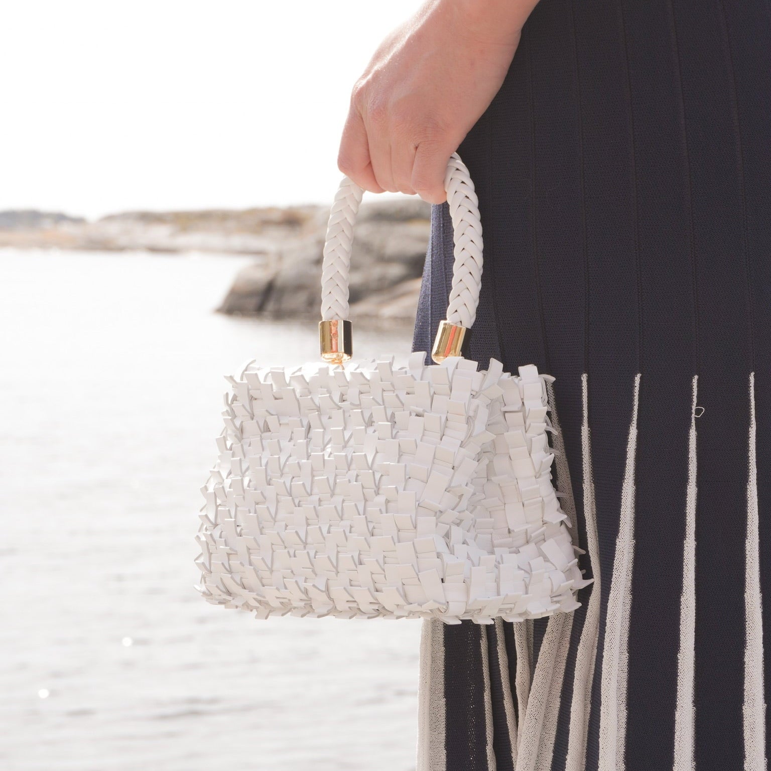 Ribichini Ribimini White håndflettet skinnveske med flettet håndtak, holdt ved vannet og stylet med mørk blå og hvit sommerkjole.