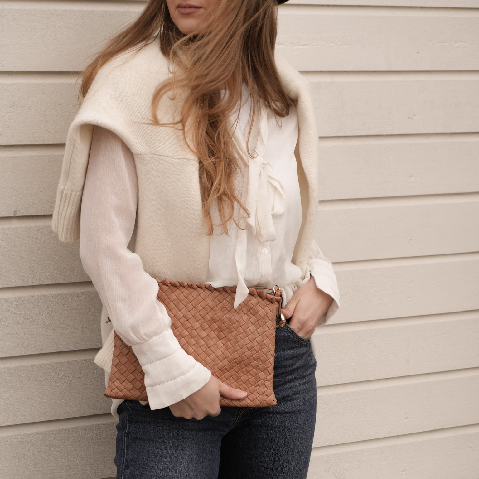 Ribichini Woodstock Clutch Cognac håndflettet skinnveske stylet som clutch, båret med hvit bluse, jeans og kremhvit genser over skuldrene.