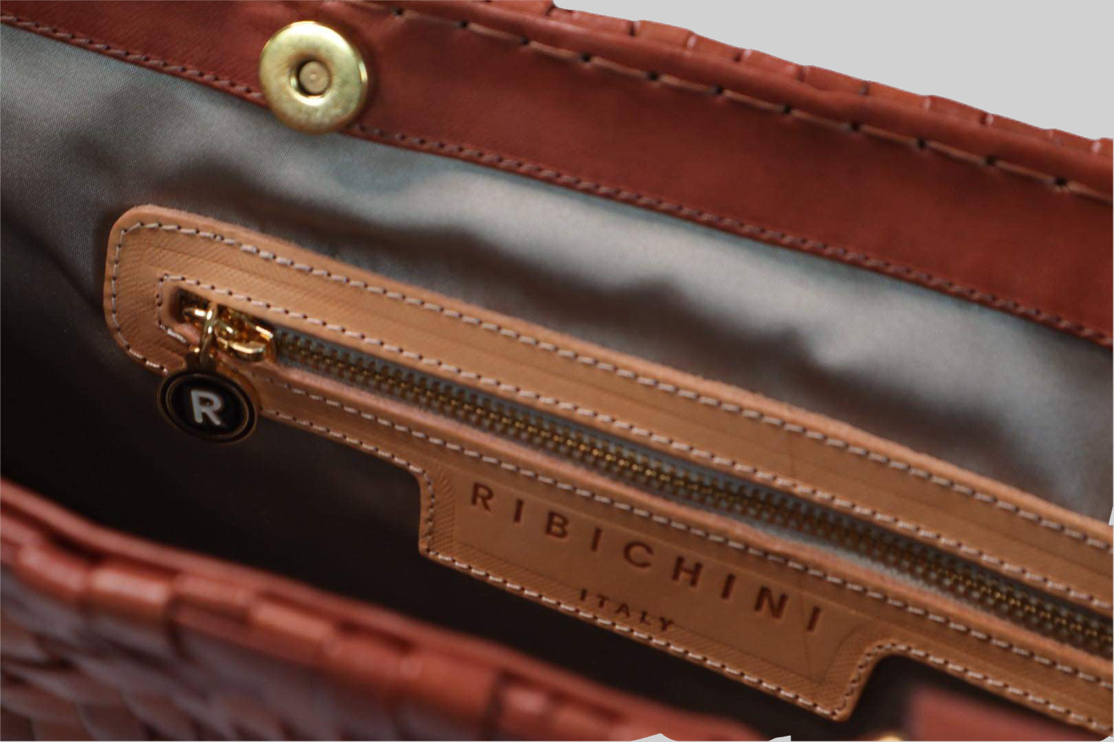 Bildet gir et detaljert inntrykk av innsiden av Ribichini Safari-vesken i cognacfarge. Med en gullfarget glidelåslomme og den ikoniske R-logoen, kombinerer den praktisk funksjonalitet med elegant design.